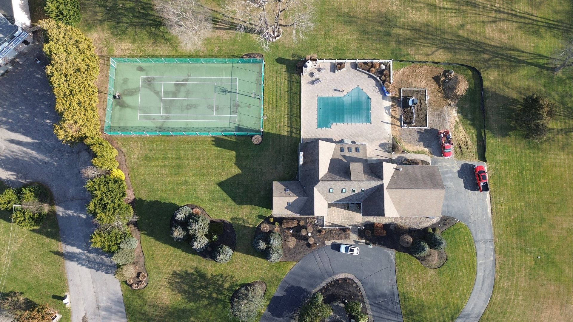Aerial view: House with pool, tennis court, and driveway on a grassy property.