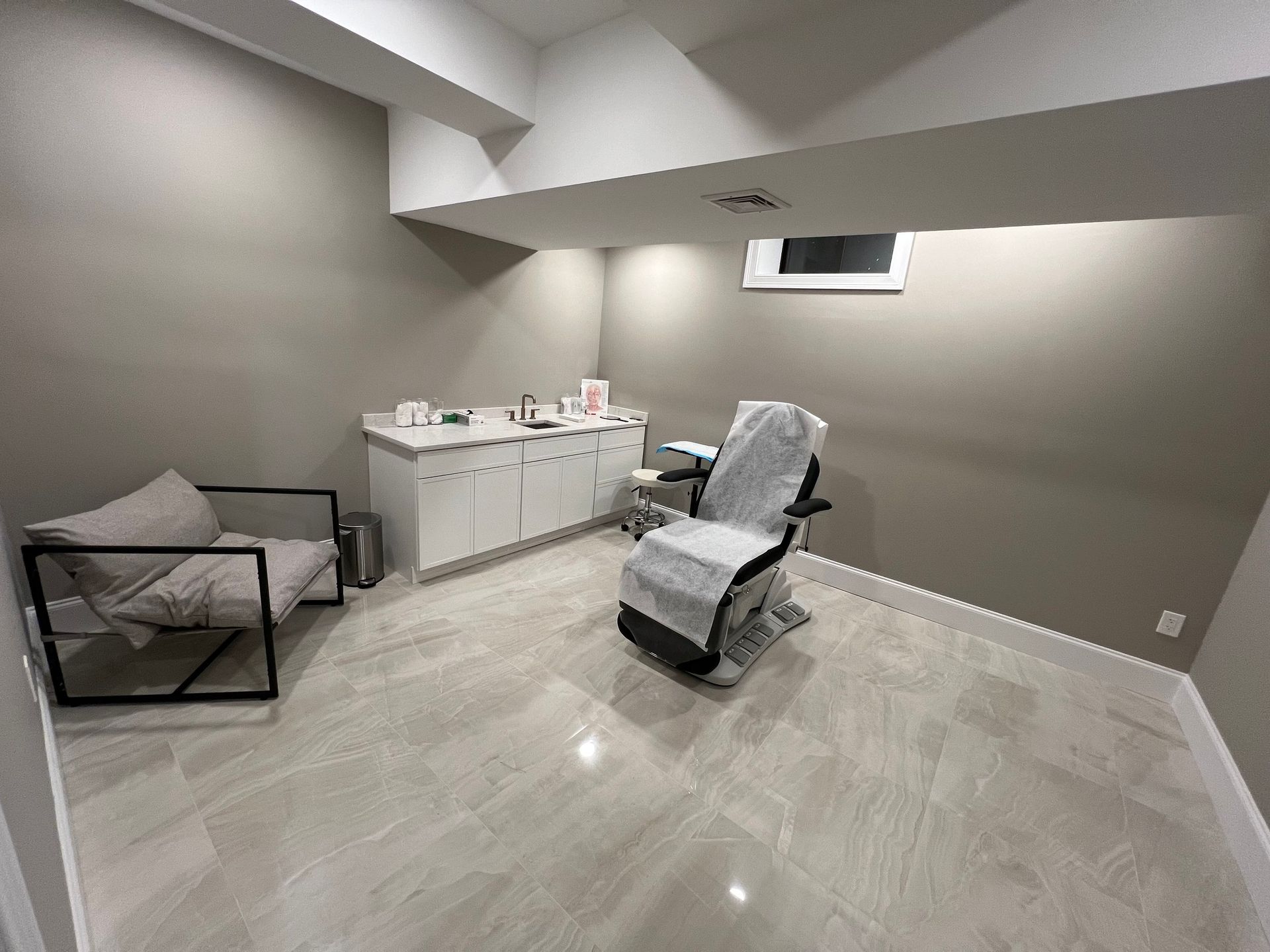 Treatment room with chair, cabinets, and a waiting area chair. Gray walls and flooring.