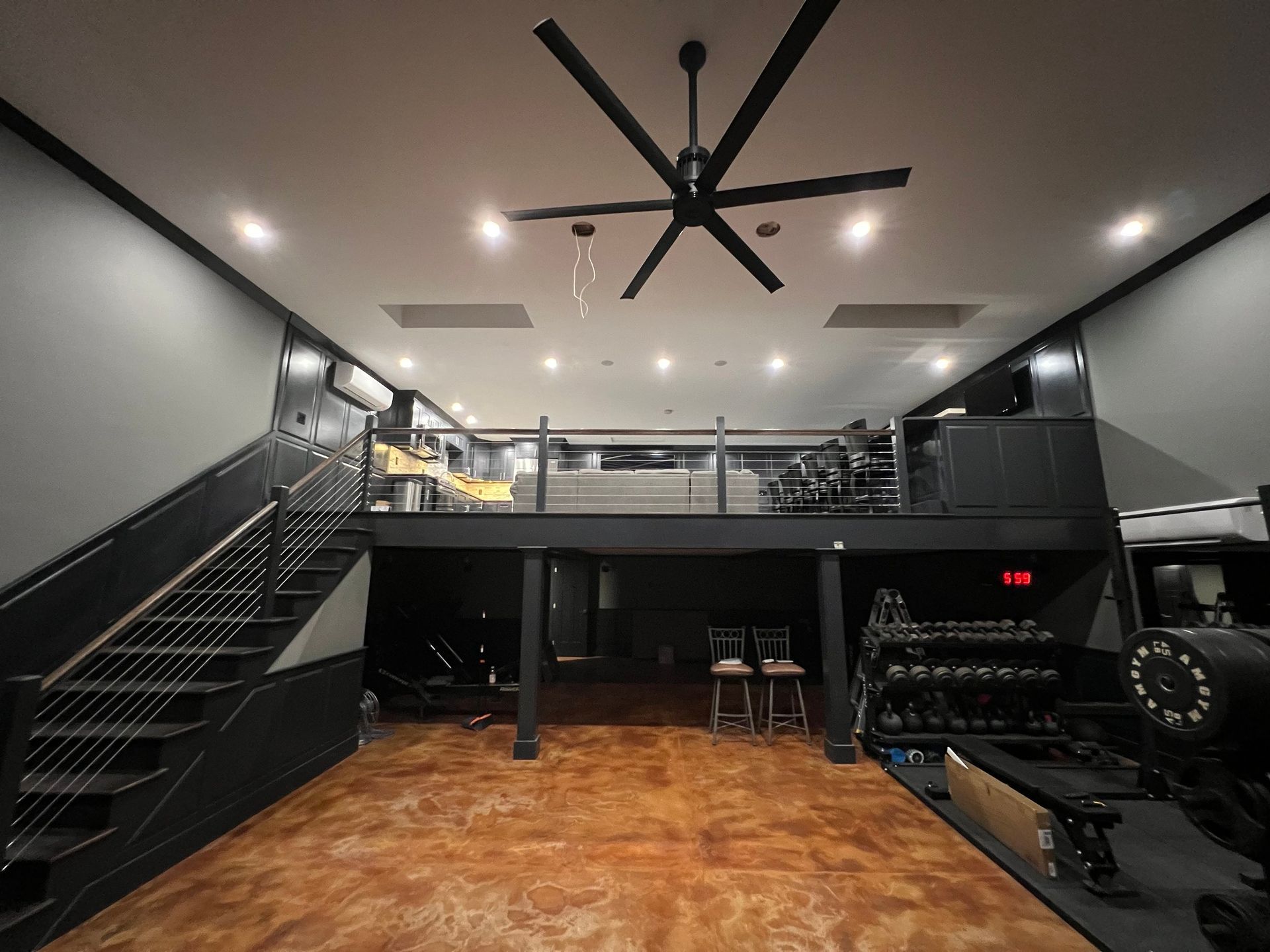 Gym with a loft, dark walls, metallic floor, weights, a large fan, and a staircase.