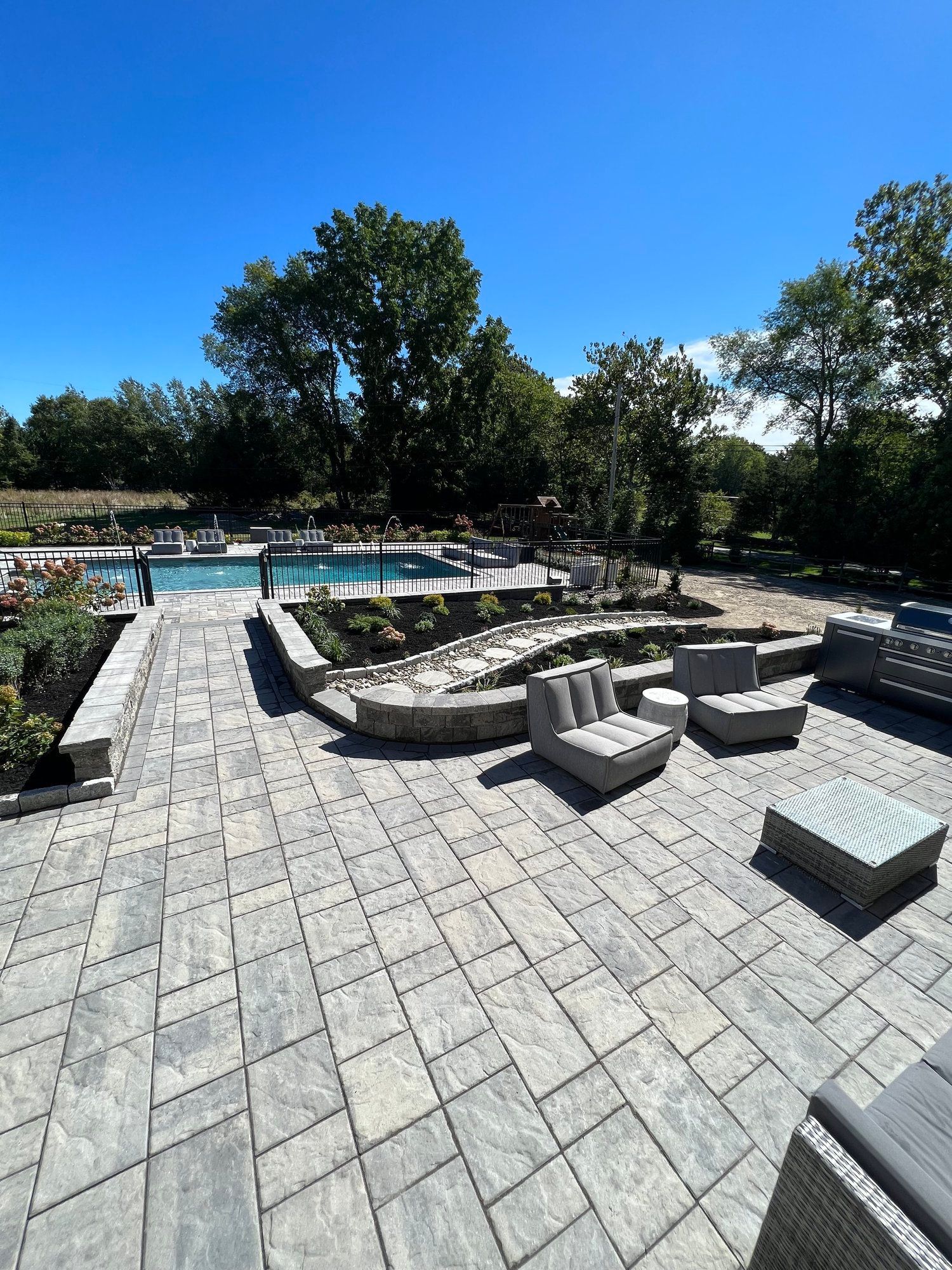Patio with gray pavers, lounge seating, and a pool. Trees and blue sky in the background.