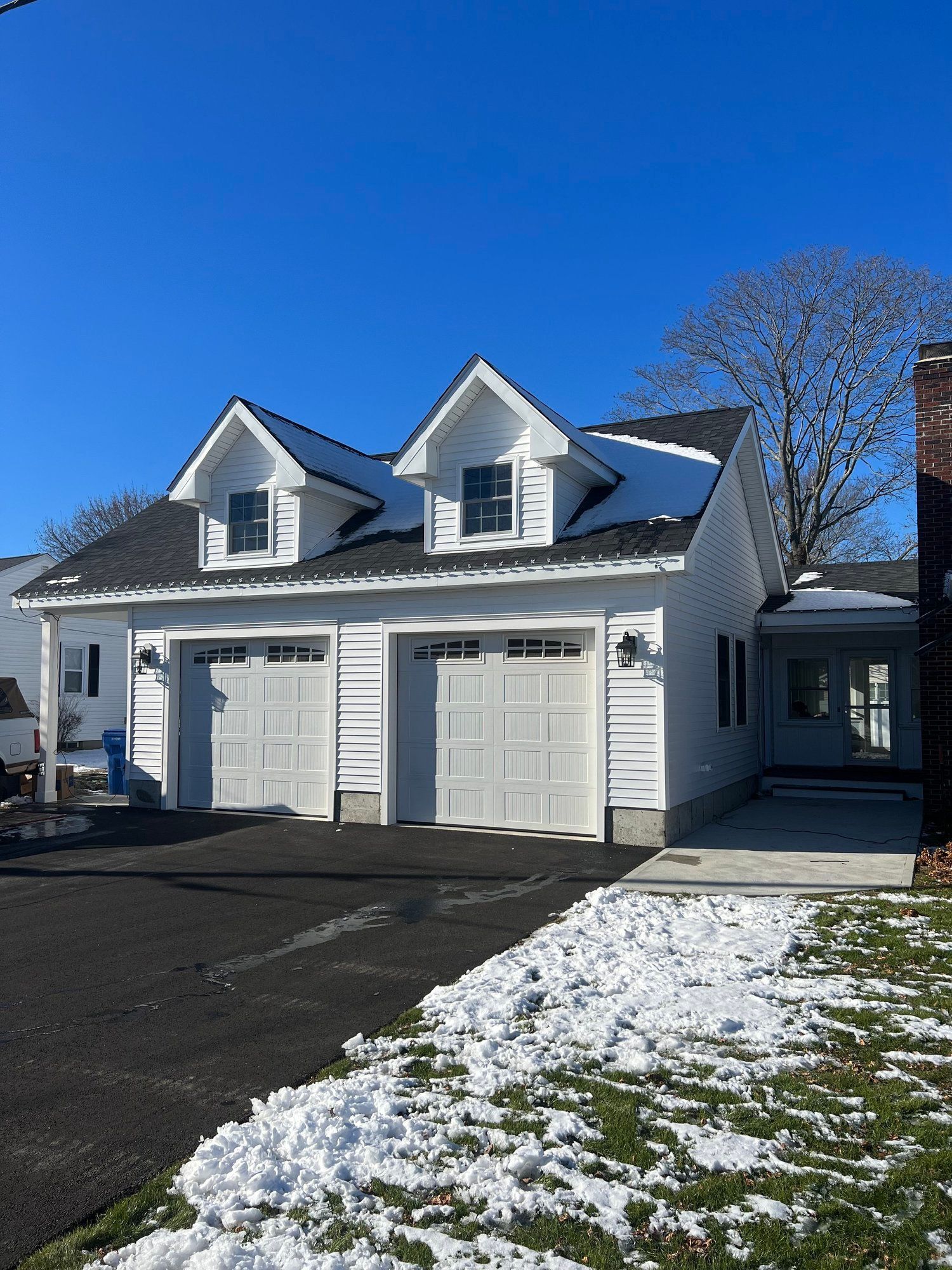 White two-car garage with two dormers on a sunny day with patches of snow on the ground.