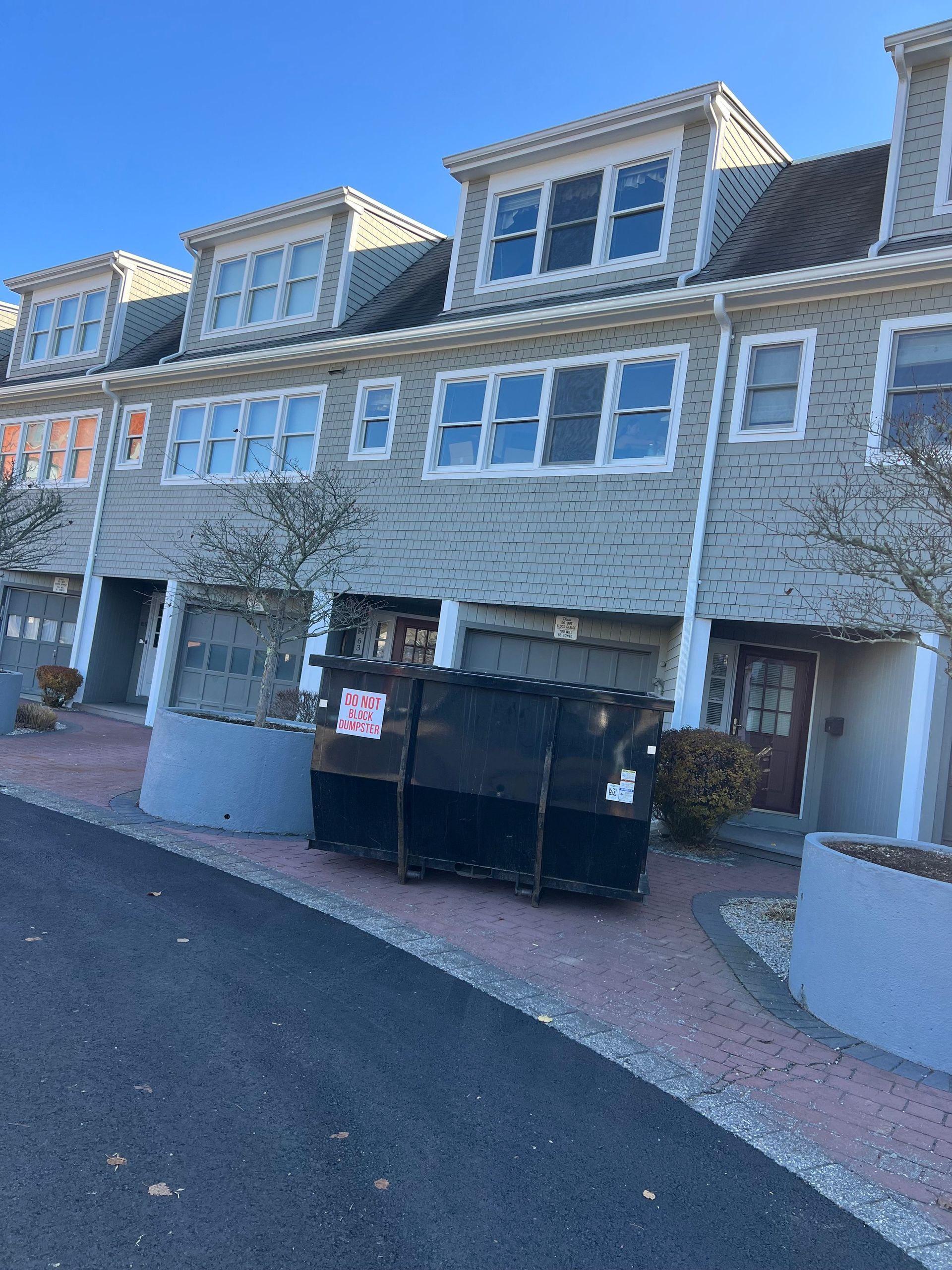 Row of townhouses with gray siding, dormers, and a large dumpster in front.