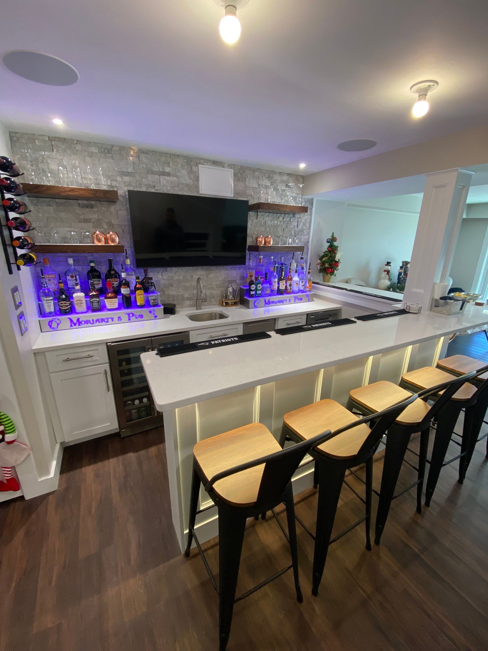 Basement bar with white countertops, black stools, liquor bottles, and a TV. Backlit with blue lights.
