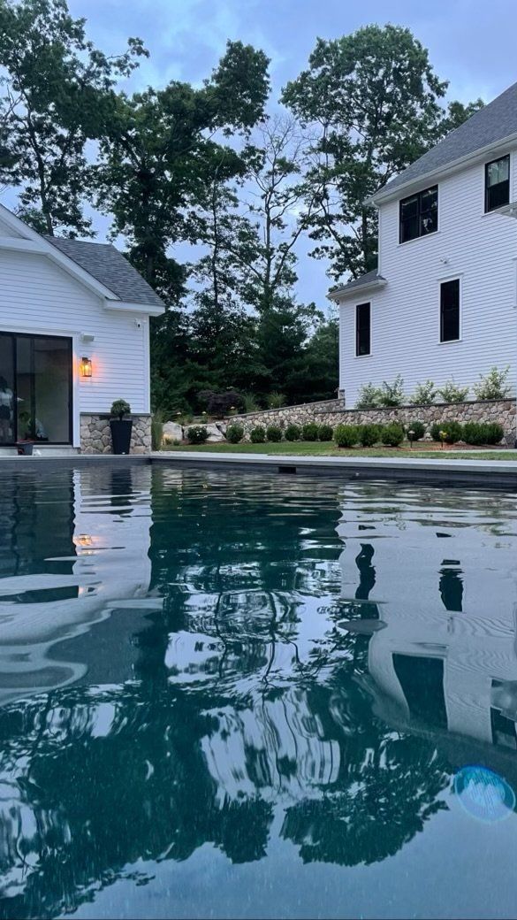 Pool reflecting white house and building with trees in the background. Cloudy sky.