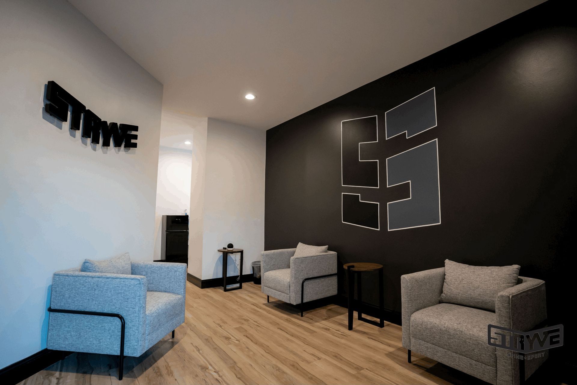 Lounge area with light wood floors, grey armchairs, black accent wall with logo, and 