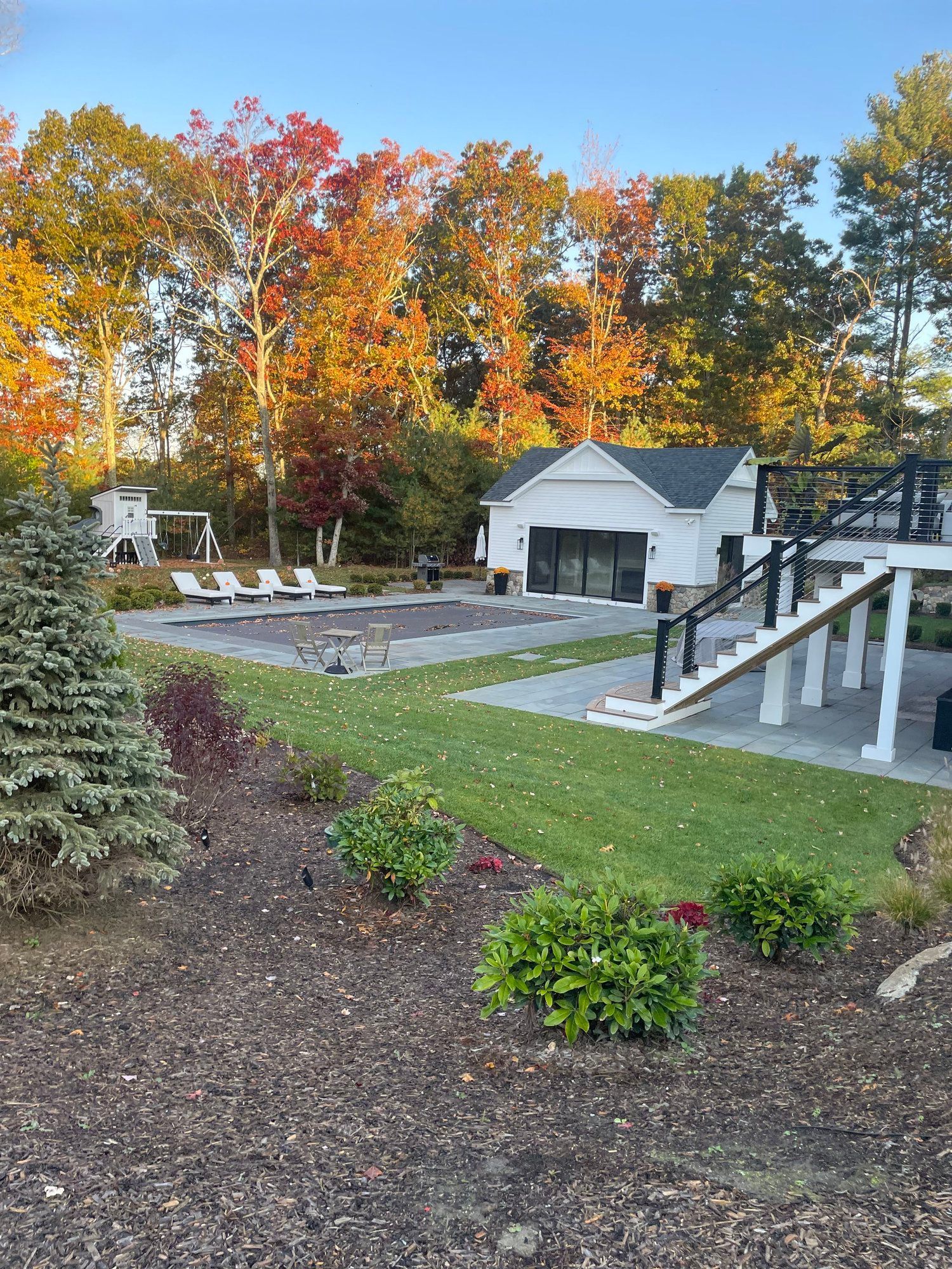 Backyard pool with white buildings, trees with fall colors, and grassy areas.