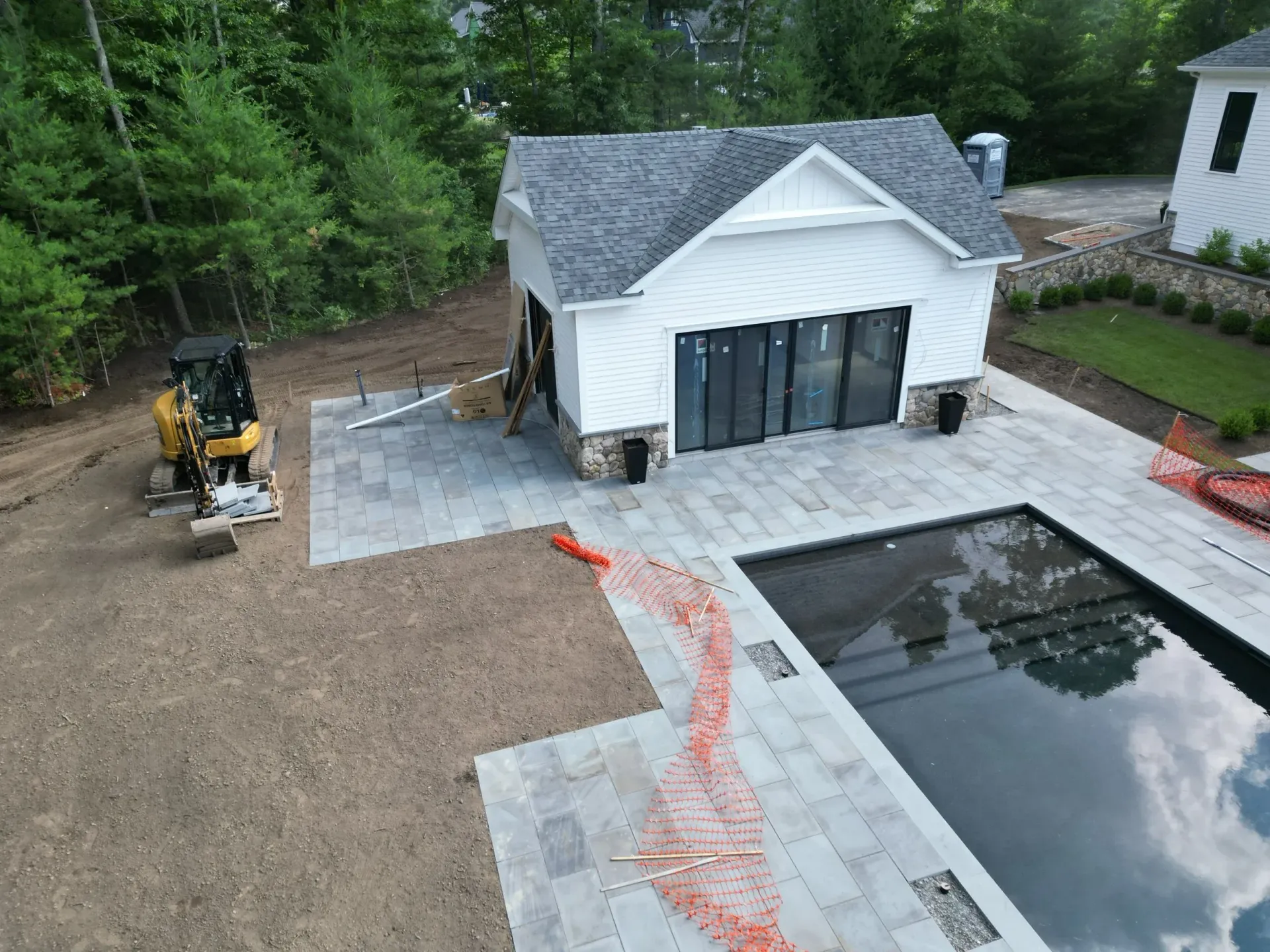 Construction site with a pool, small white building with black doors, and a small excavator.