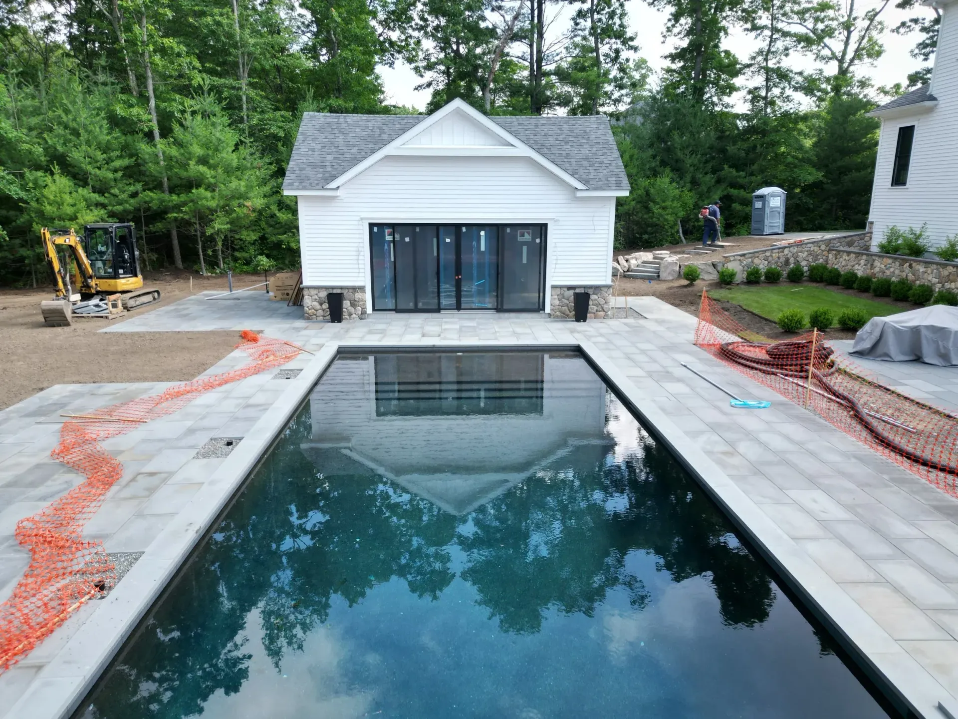 Pool with white cabana, surrounded by gray pavers. Trees in background, construction materials visible.