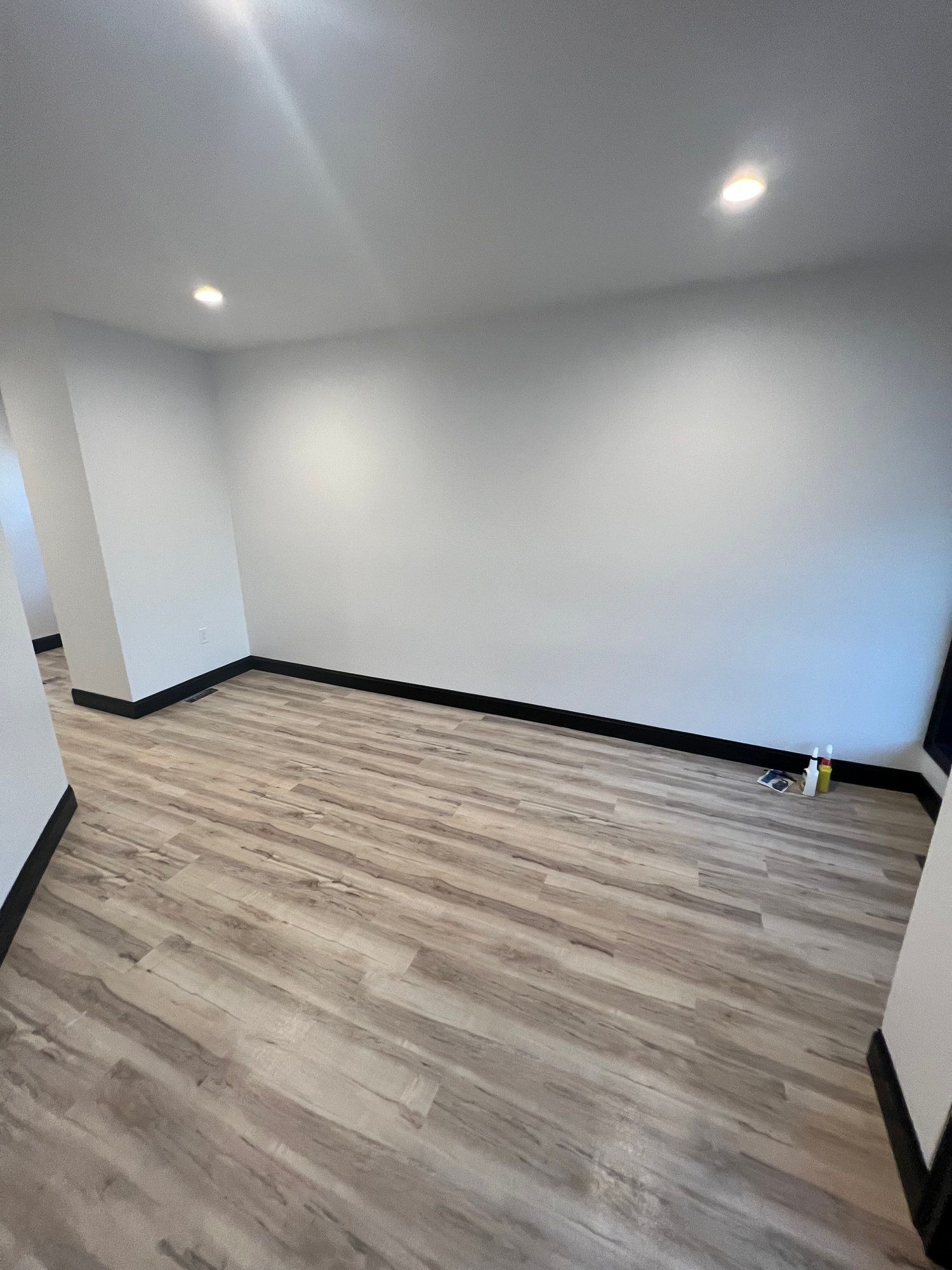 Empty room with light wood-look flooring, white walls, and black baseboards. Recessed lighting.