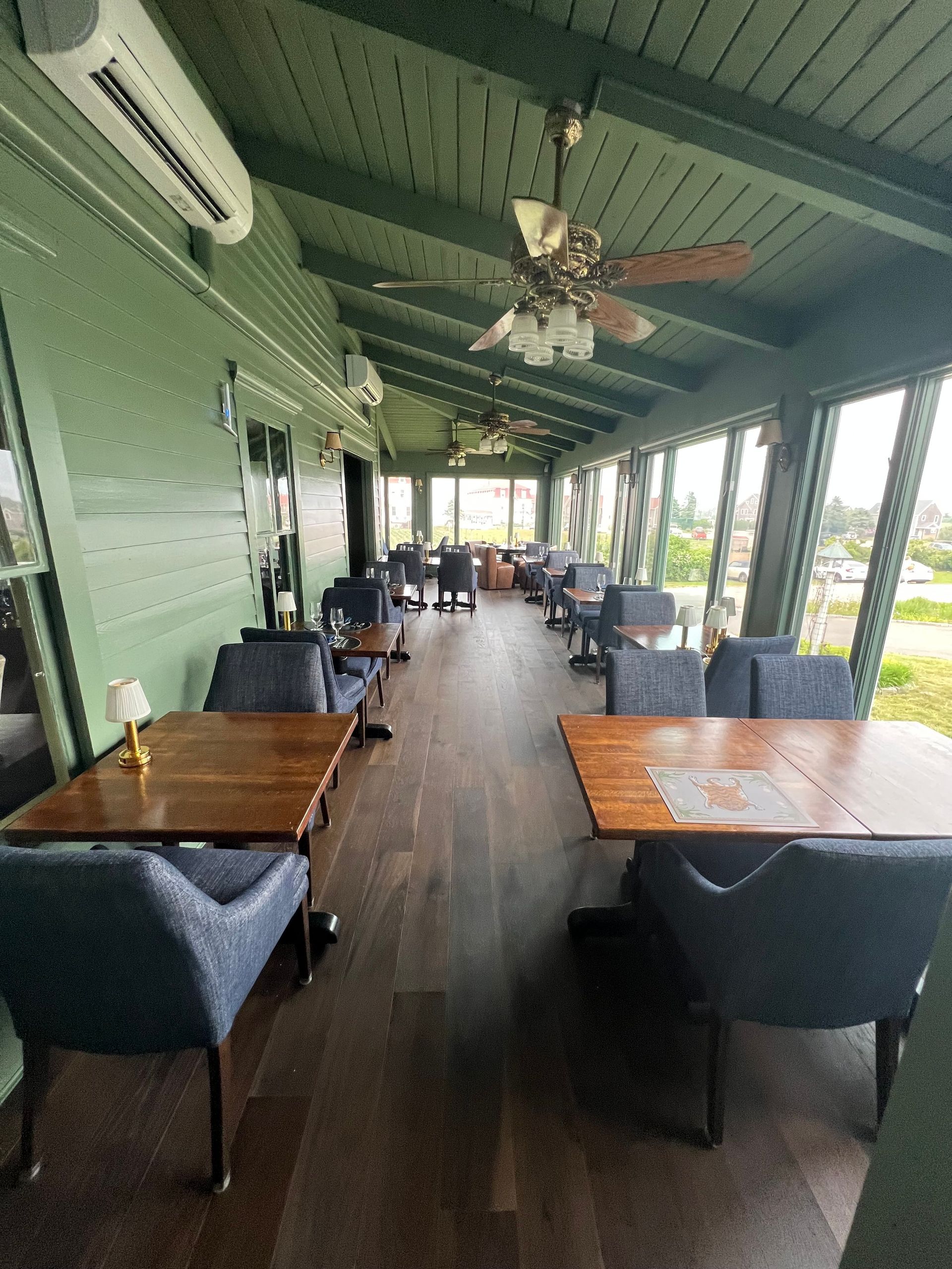 Green-walled cafe with wooden floors and tables, blue chairs, and a ceiling fan, overlooking a view.