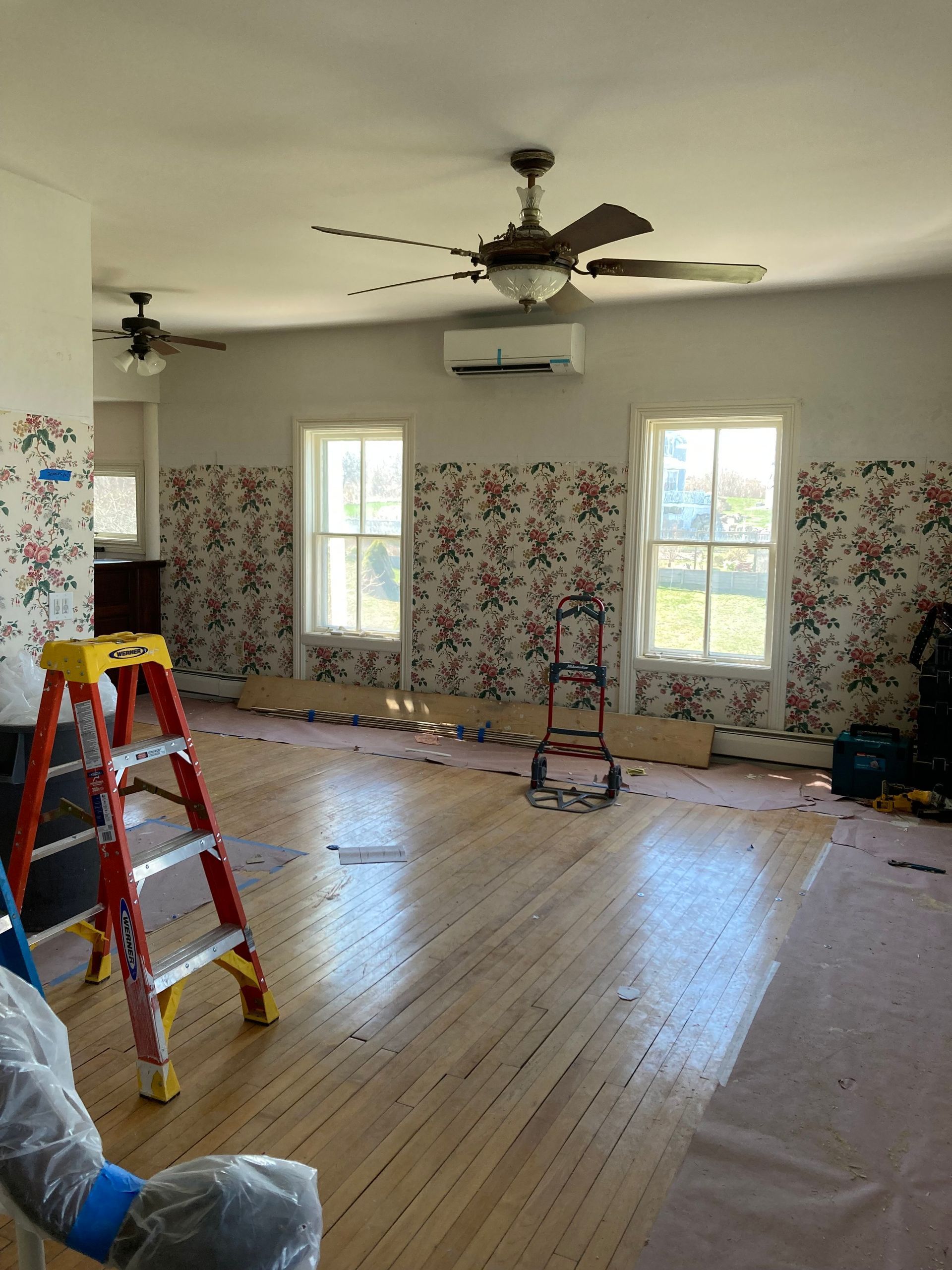 Room interior with floral wallpaper, two windows, two ceiling fans, ladder, and wood flooring.