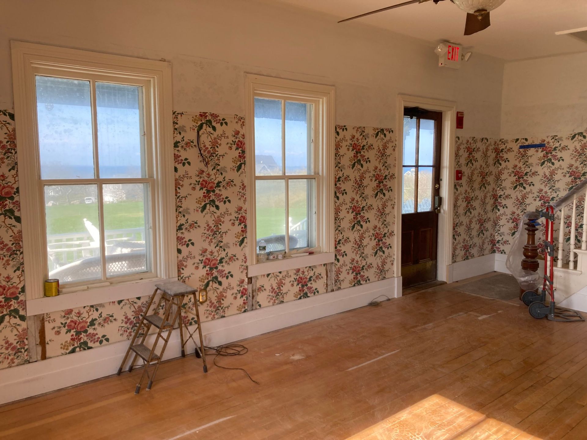 Interior with windows, floral wallpaper, wooden floor, and a staircase. Light streams in; a stepladder rests near windows.