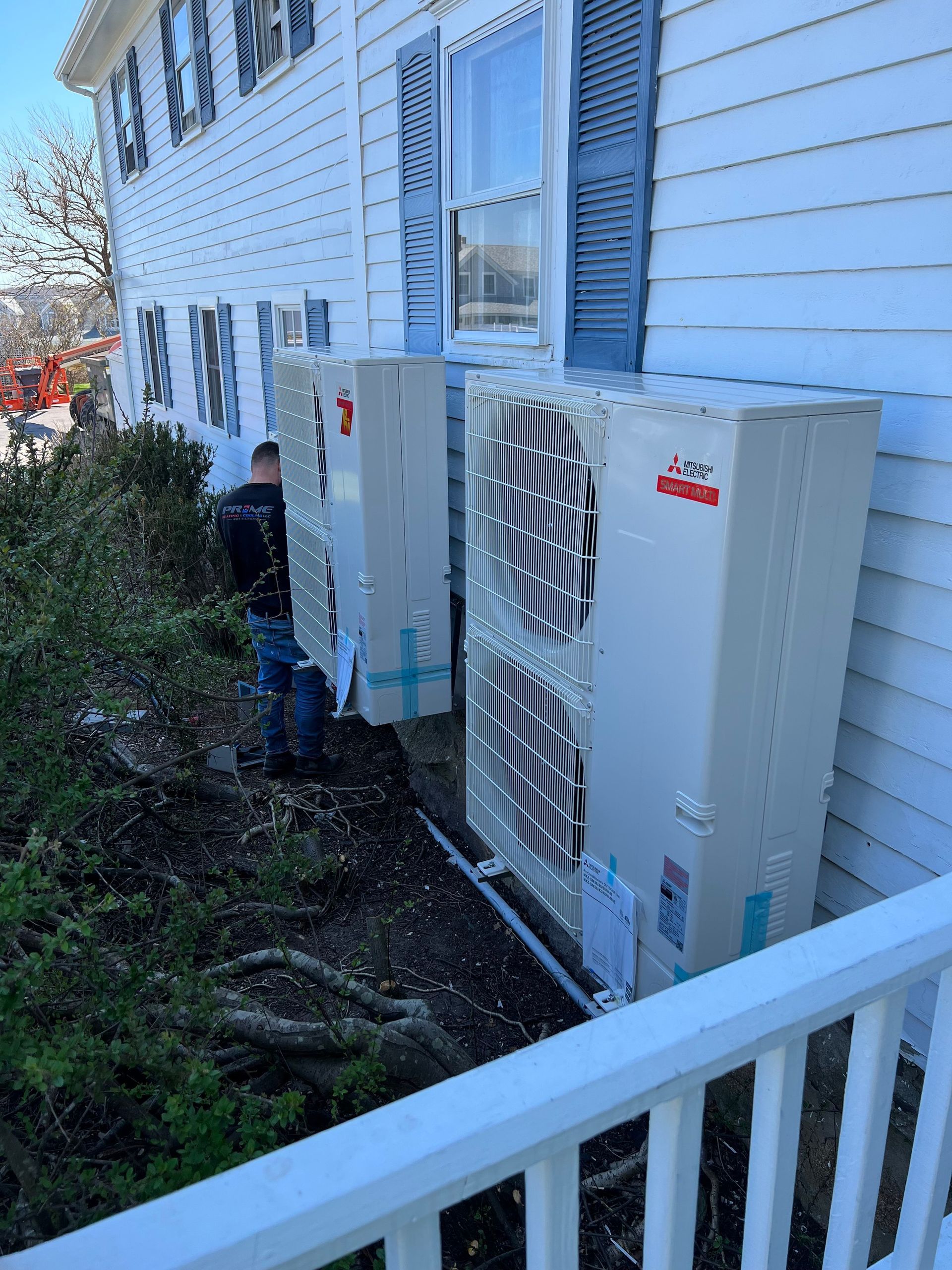Two white air conditioning units installed outside a white house with blue shutters. Workers nearby.