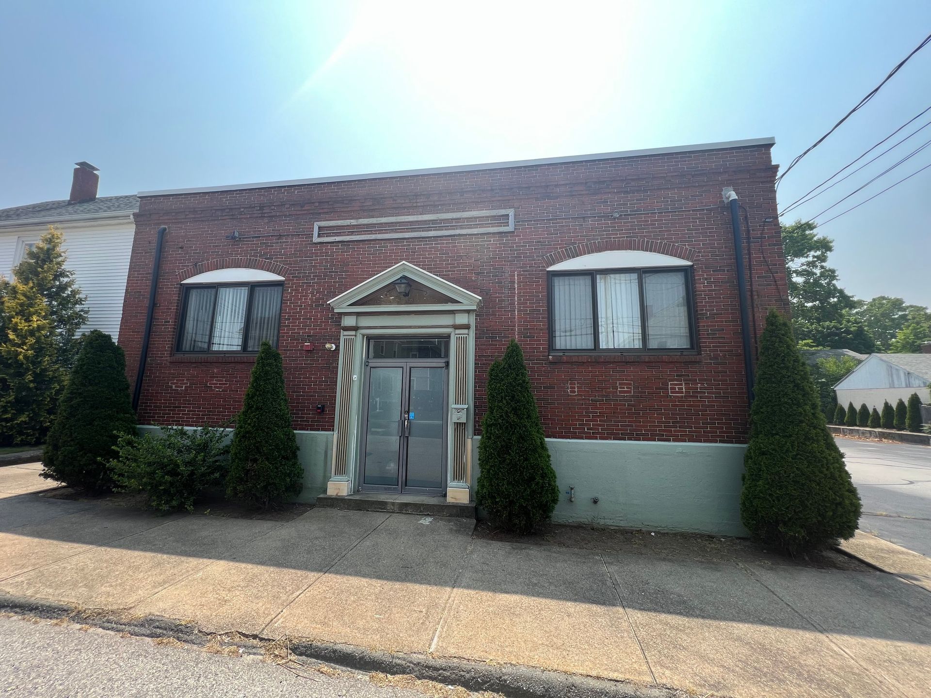 Red brick building with two windows, a decorative doorway, and small evergreen trees.