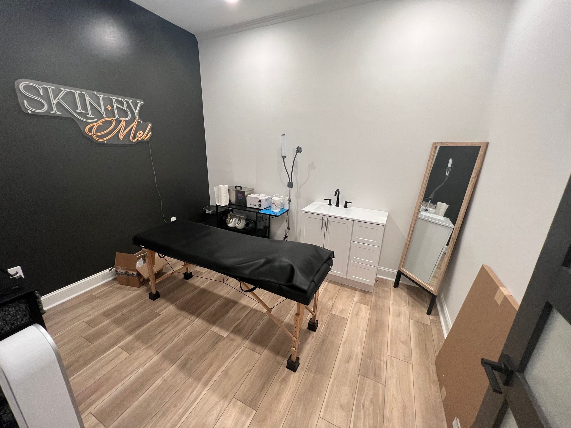 A room with a massage table, cabinet, and mirror.  The wall behind the table is dark with a neon sign that says 