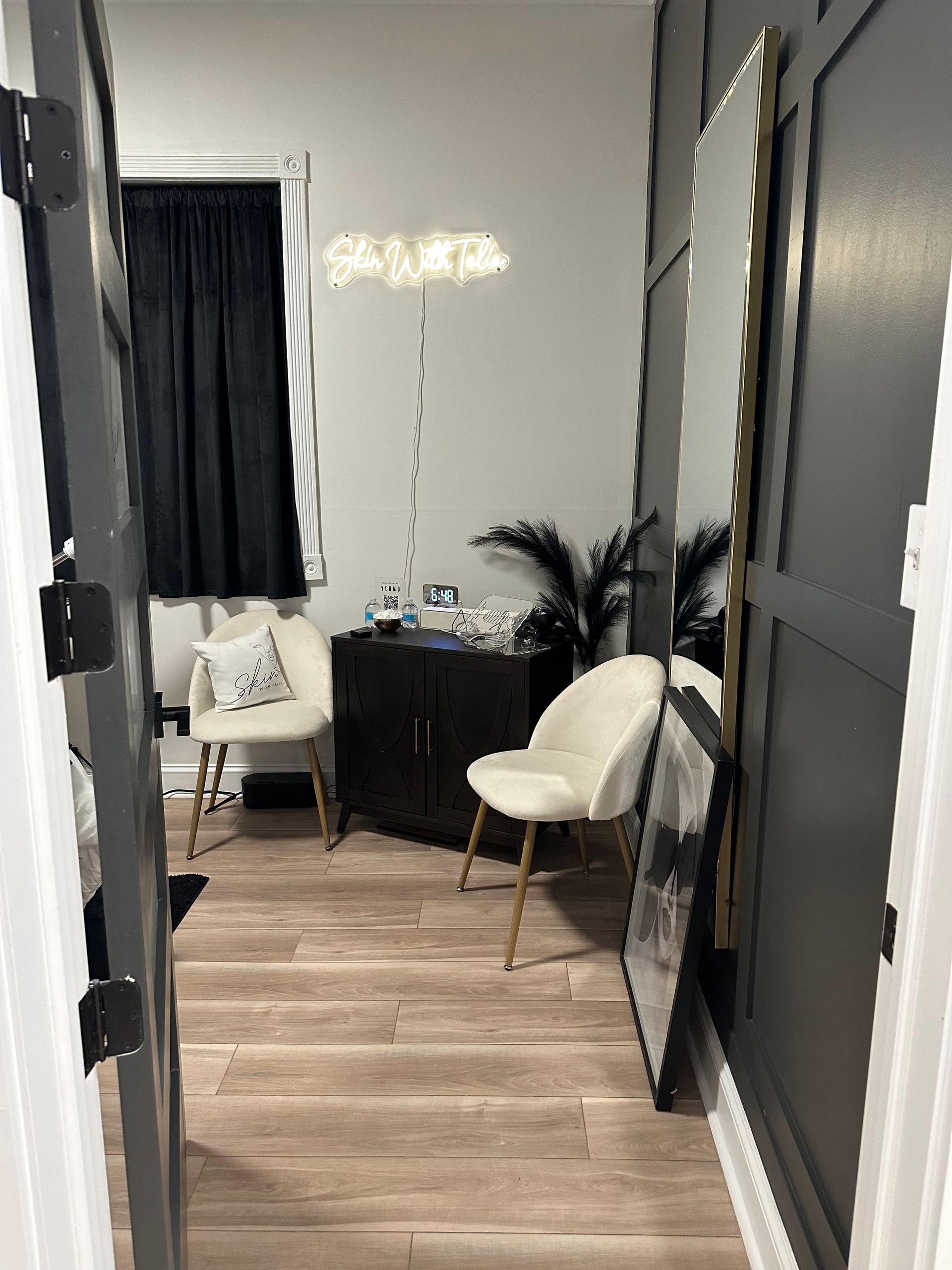 A small room with light wood floors, two white chairs, a dark cabinet, and a large gold mirror.