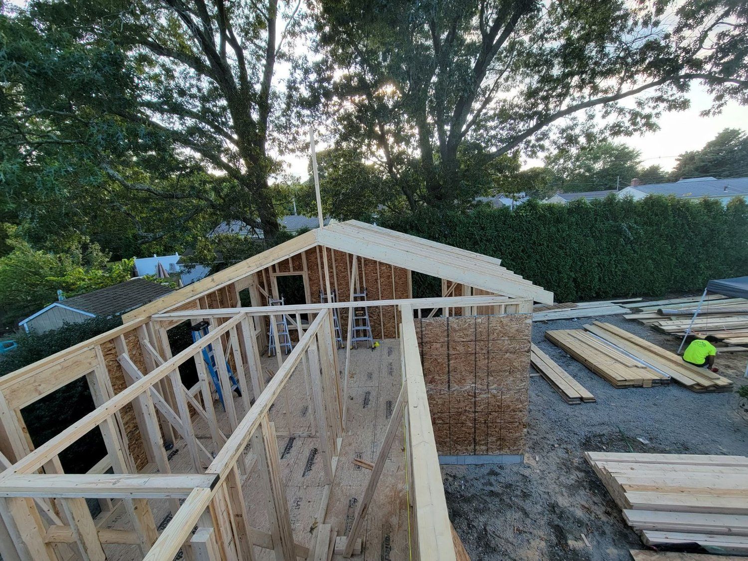 Wooden frame of a building under construction, exterior view. Person near lumber. Trees in the background.