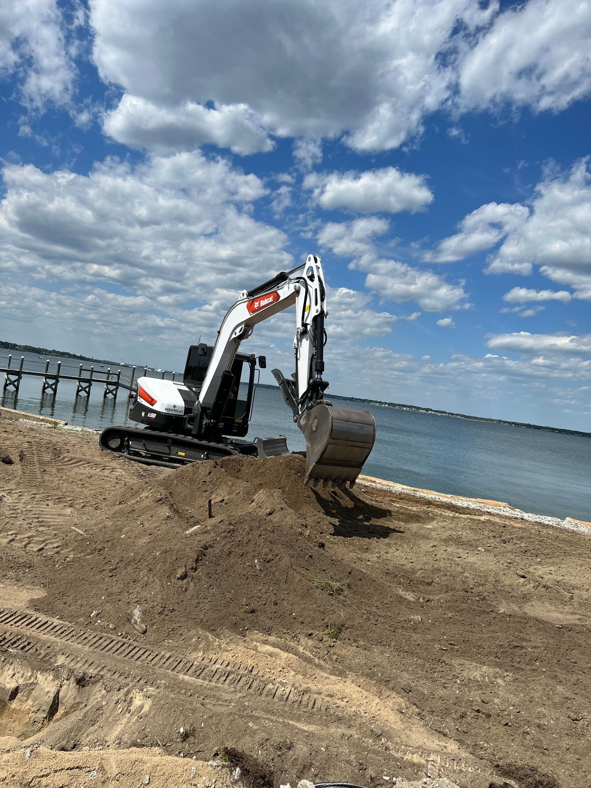 Bobcat excavator digging on a sandy beach, with water and a cloudy sky in the background.