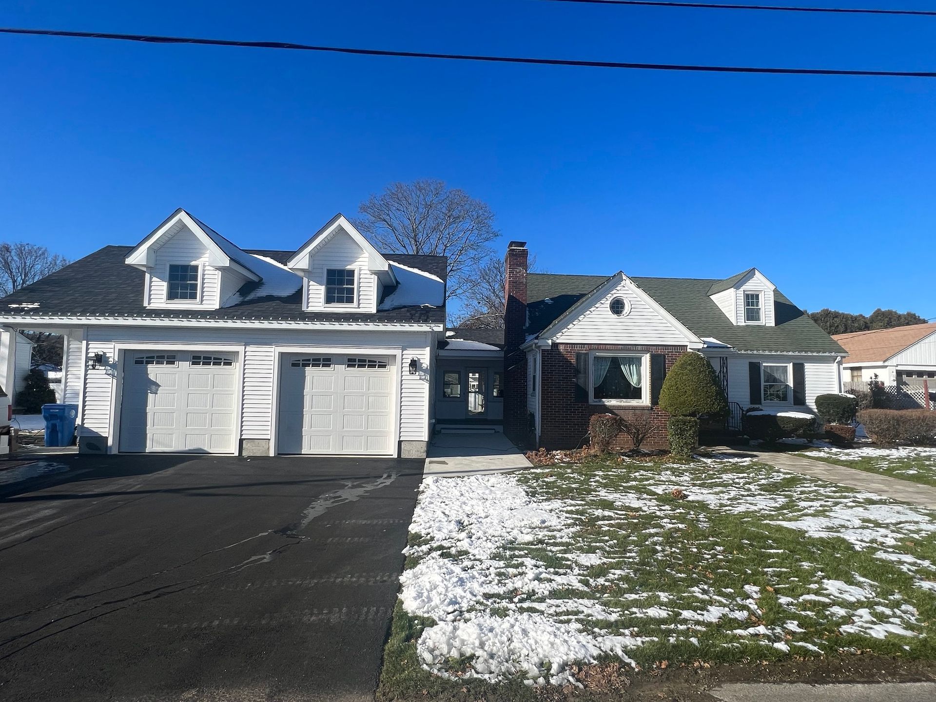 Two houses side-by-side on a sunny day. One has a white facade and attached garage. The other has a brick facade and a green roof. Snow on the ground.