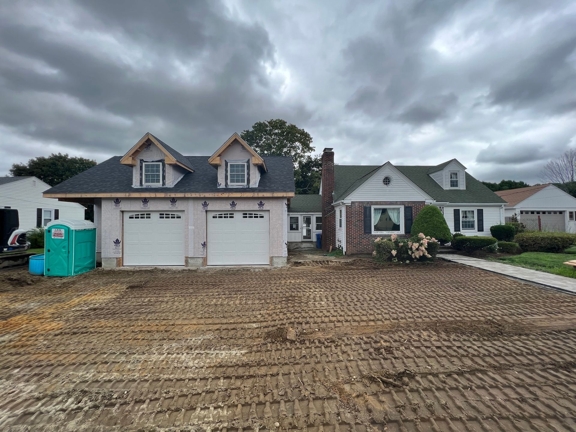 A house with a newly constructed garage, dirt lot in the foreground, cloudy sky overhead.
