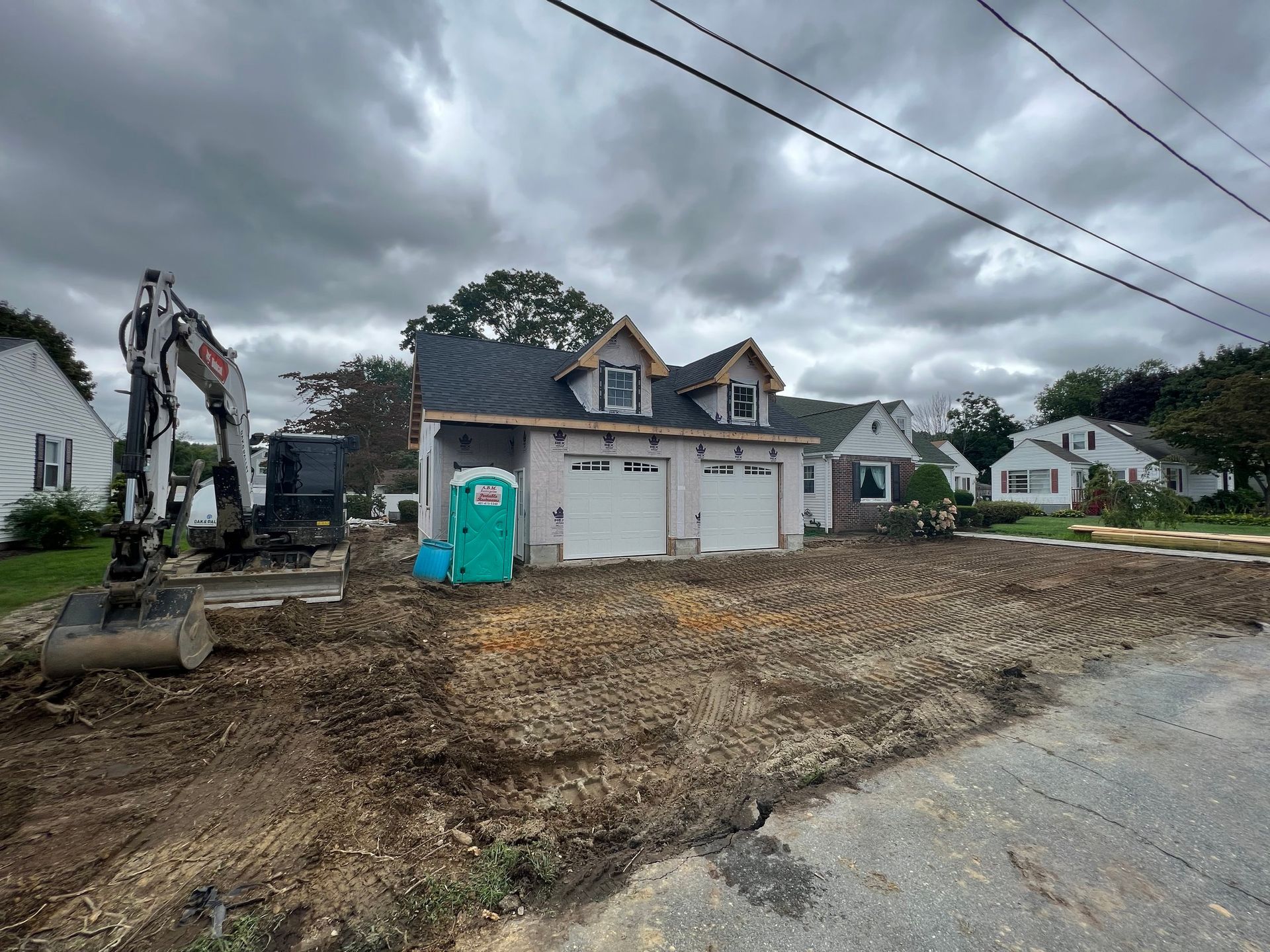 Construction site: Two-car garage under construction with an excavator and porta-potty, cloudy sky.