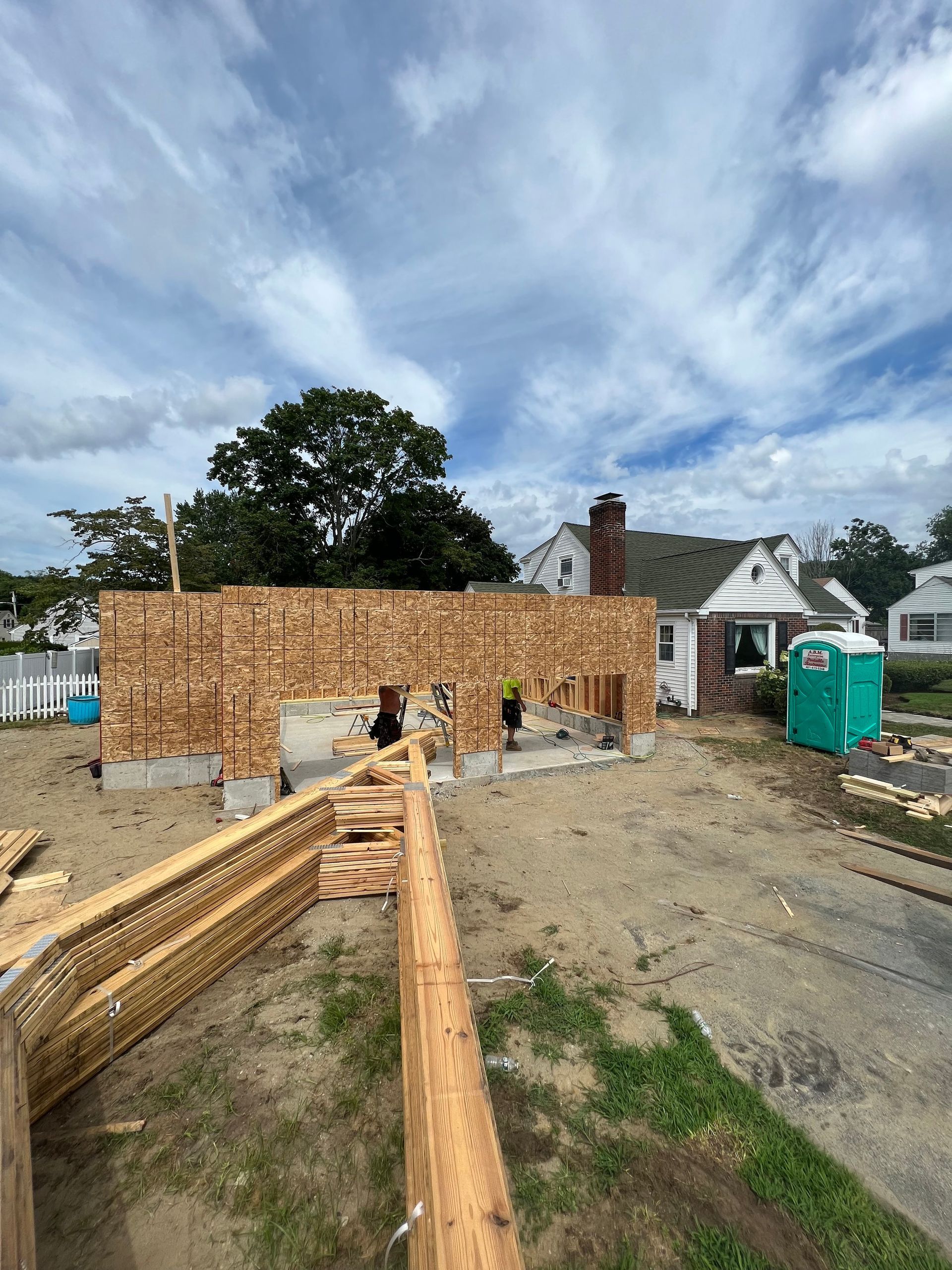 Construction site with a wooden frame, and a house in the background. Blue portable toilet.