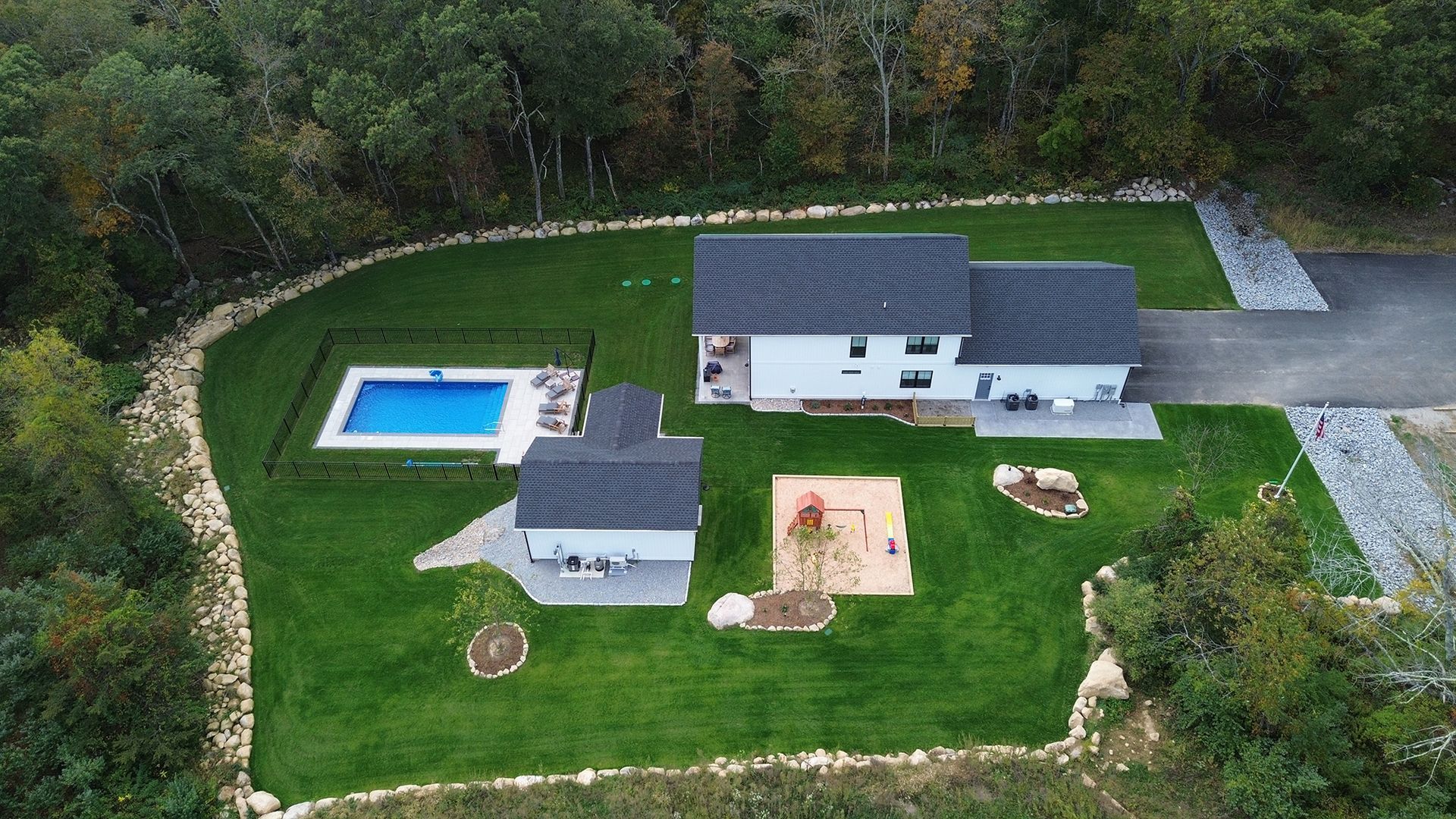 Aerial view of a white house, pool, and shed on a green lawn surrounded by trees and a gravel driveway.