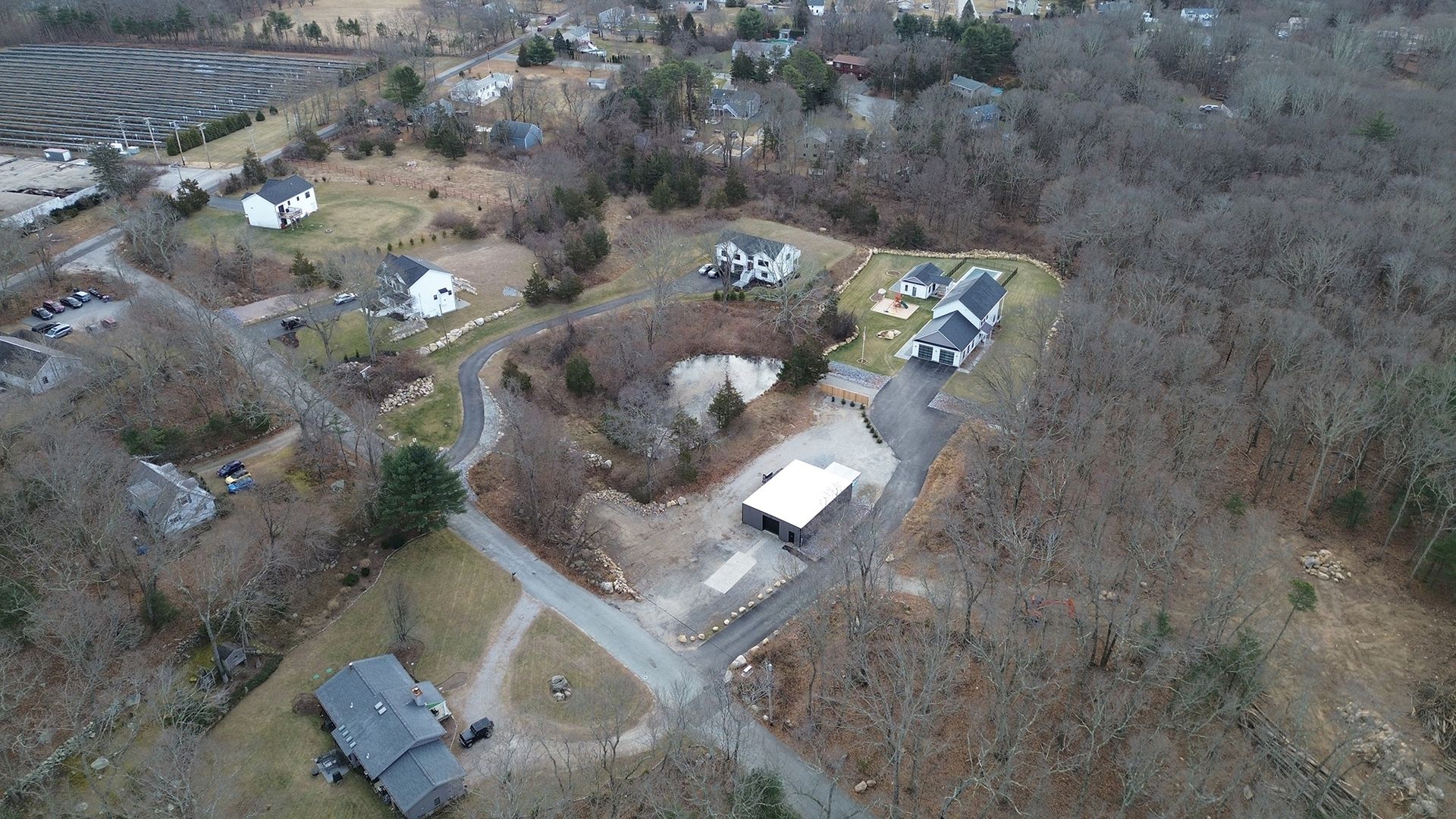 Aerial view of houses and a building in a wooded area with a paved road and a water feature.