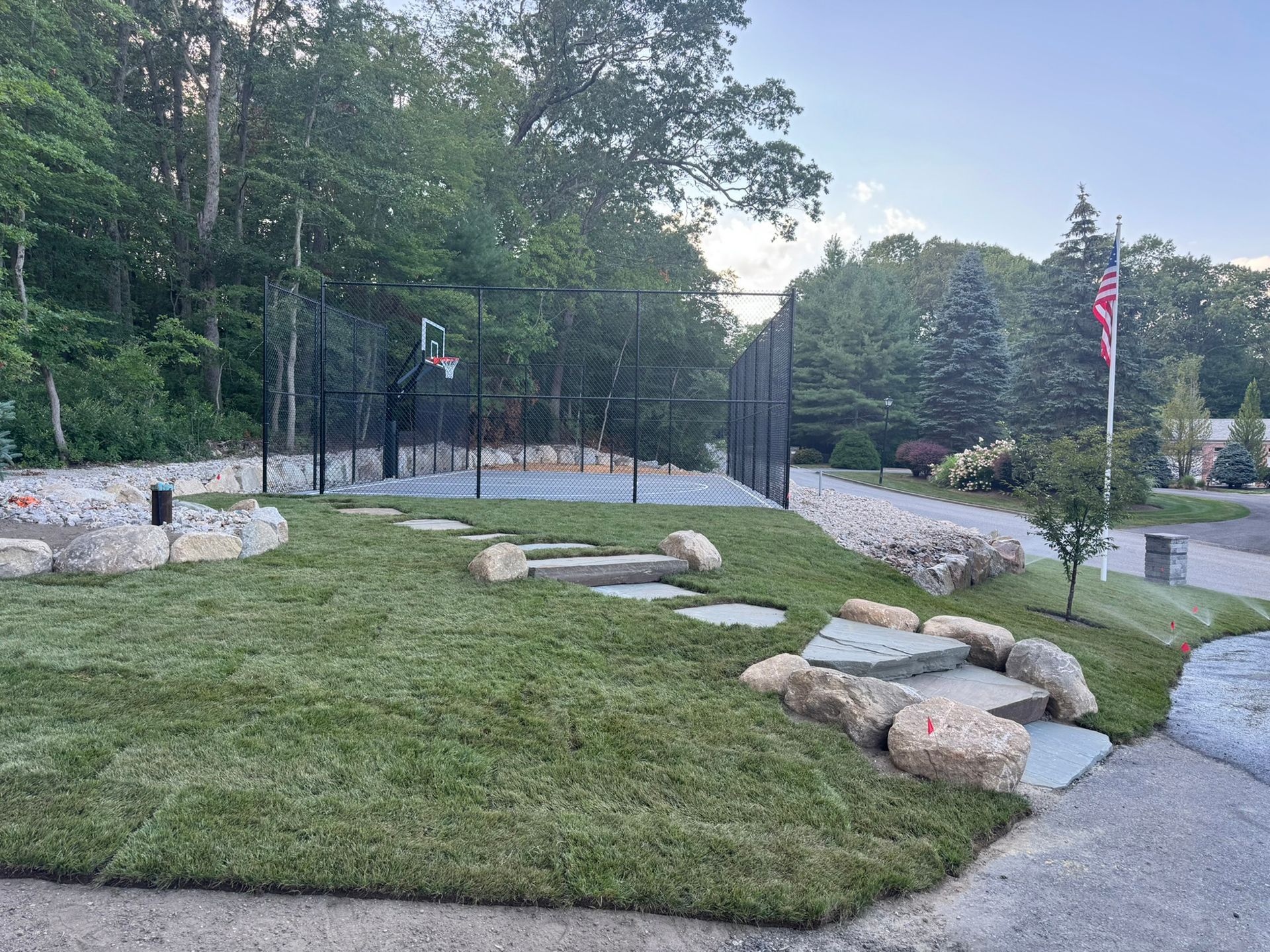Newly landscaped basketball court with black fencing, surrounded by grass and rocks. American flag waves in background.