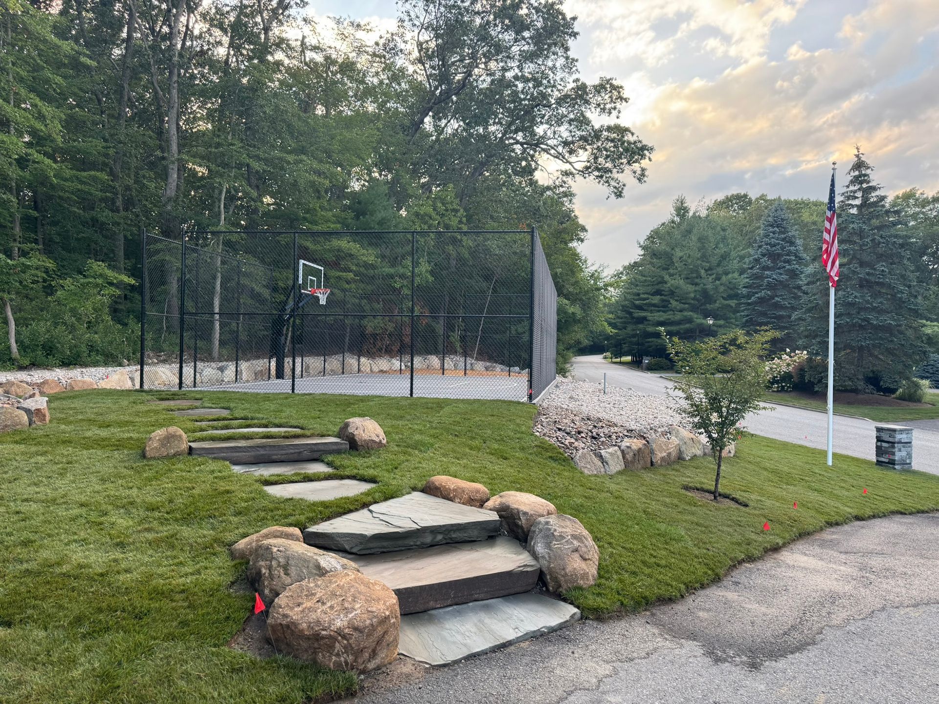 Stone steps lead to a black-fenced basketball court, lawn, and an American flag on a pole.