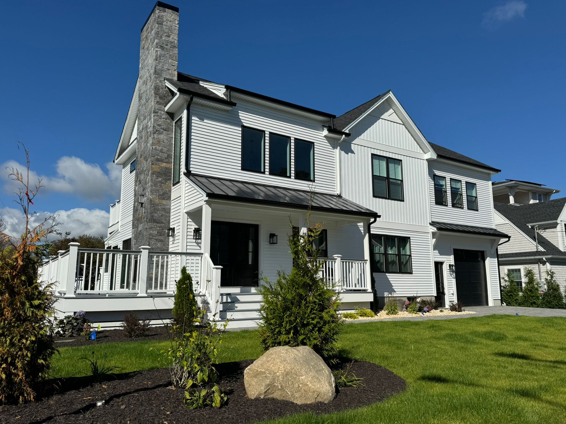 White two-story house with black trim, chimney, and roof against a blue sky; front porch and manicured lawn.