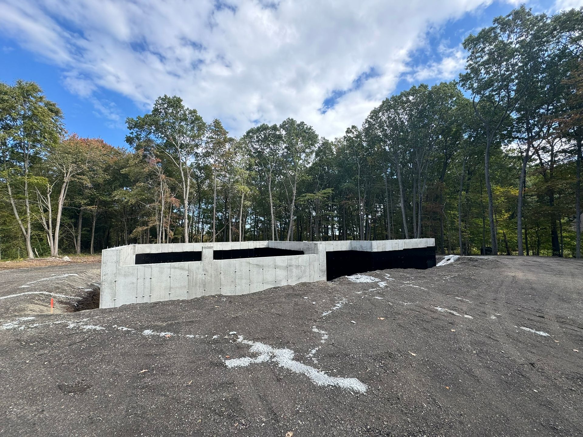 Concrete foundation of a building under construction on a dirt lot, surrounded by trees and a cloudy sky.