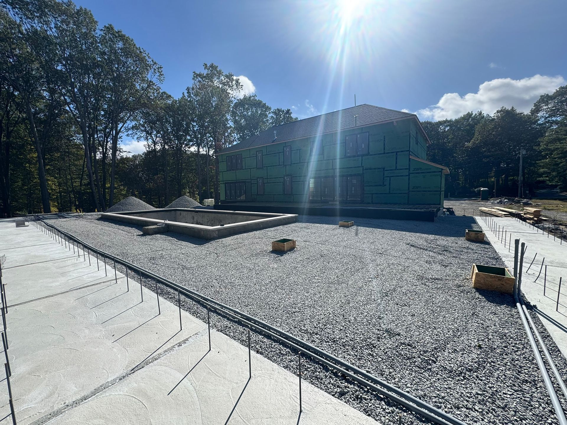 Construction site: house framed with green wrap, on gravel, with concrete foundation and walkways under a sunny sky.