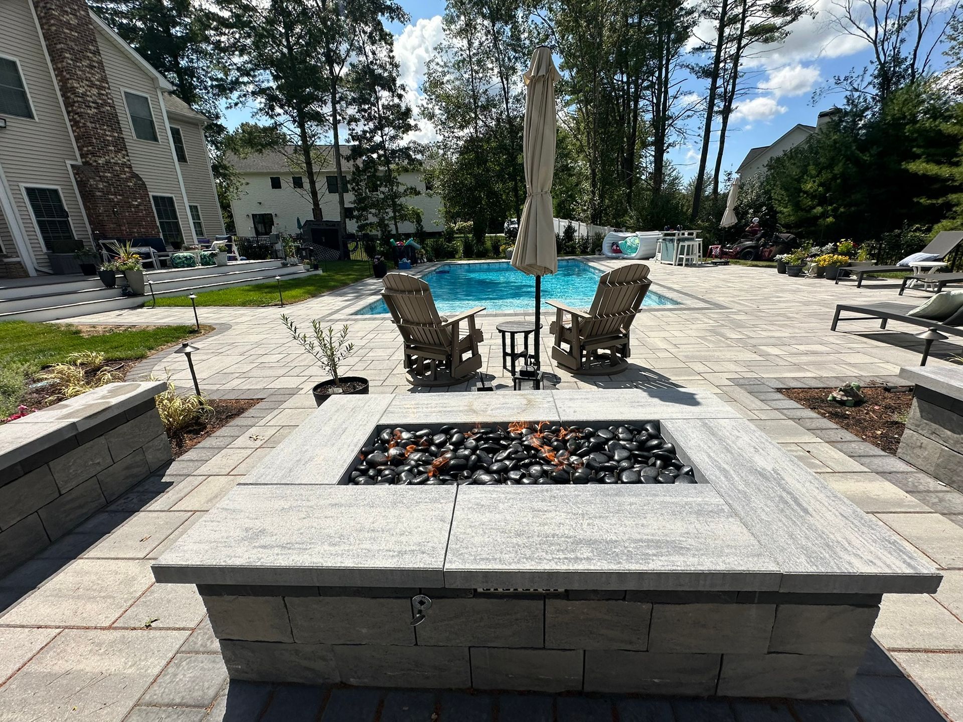 Fire pit on a patio with pool and lounge chairs in background.