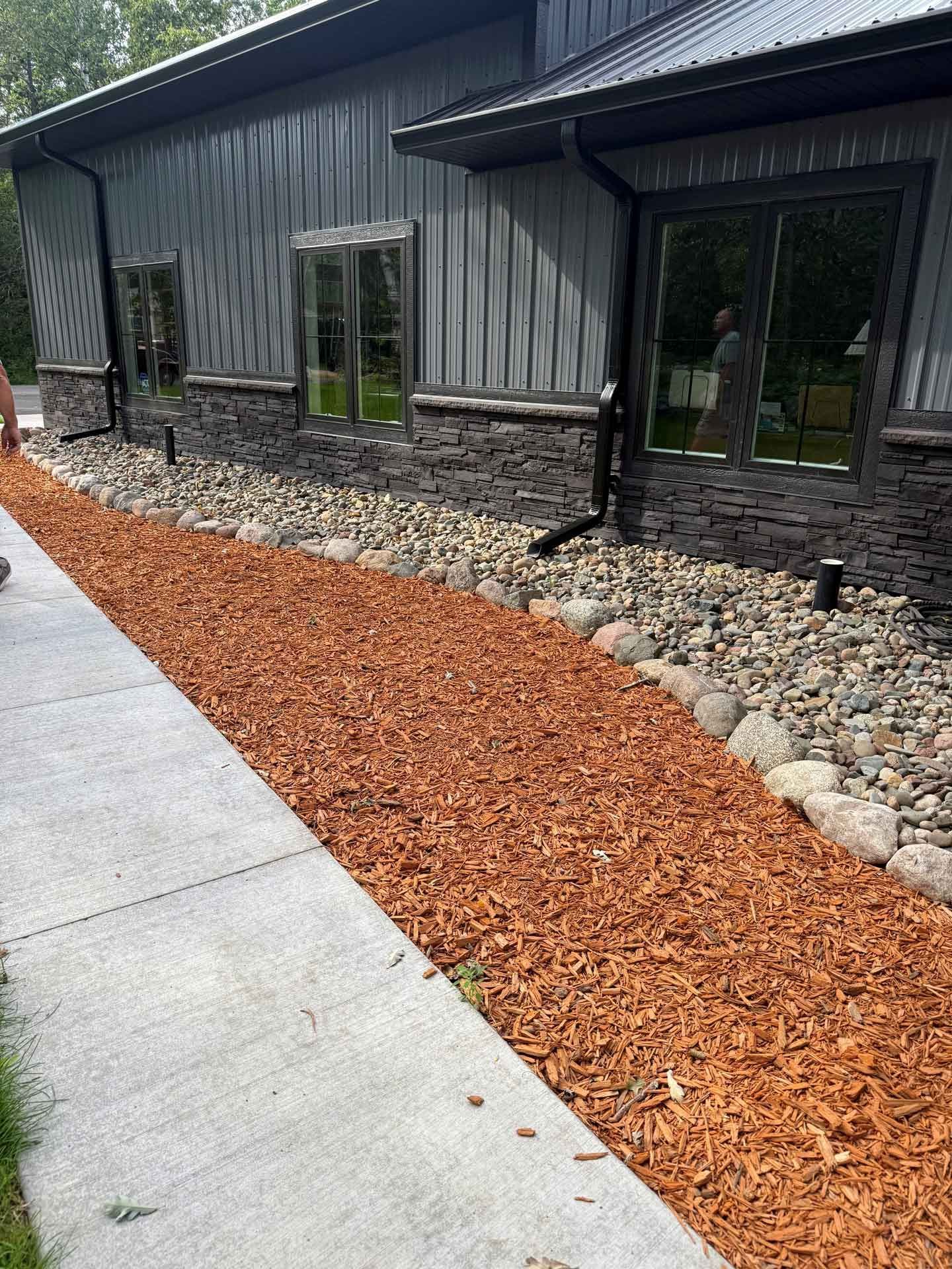 Modern building exterior with black siding, windows, stone border, and a red mulch path beside a concrete walkway