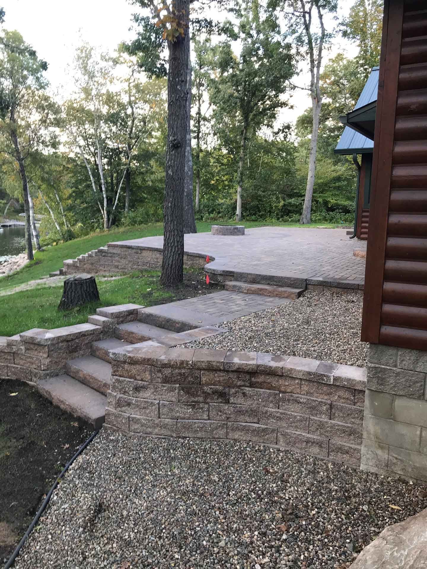 Stone steps and a gravel patio in a wooded yard beside a log cabin.