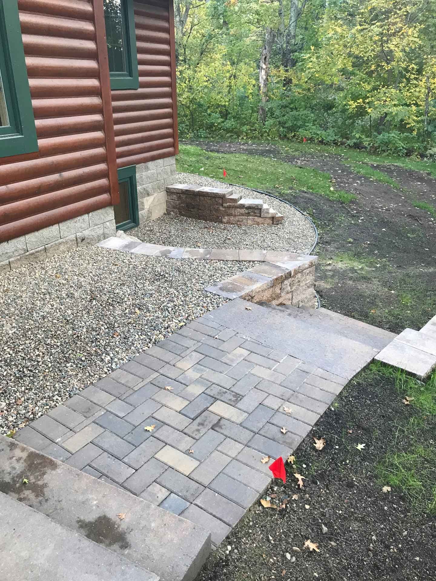 Brick and concrete walkway beside a log house, leading to a gravel area and wooded yard.
