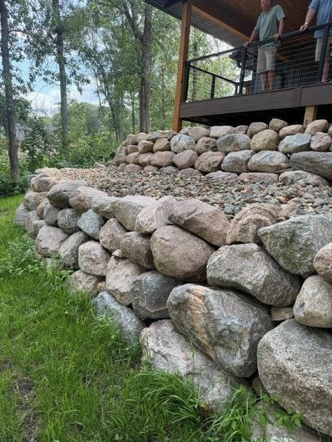 Stone retaining wall beside a grassy yard and wooden deck under trees