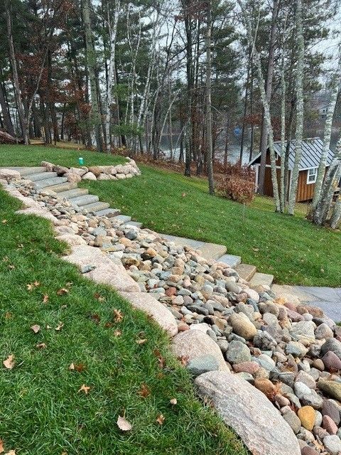 Rock-lined drainage swale beside a grassy yard with trees and a small shed in the background