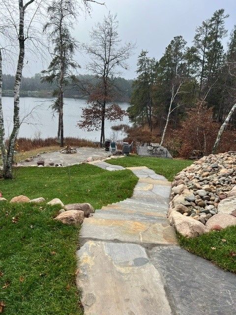 Stone path through a grassy yard leads to a lake on a cloudy day, with trees and rocks along the shore.