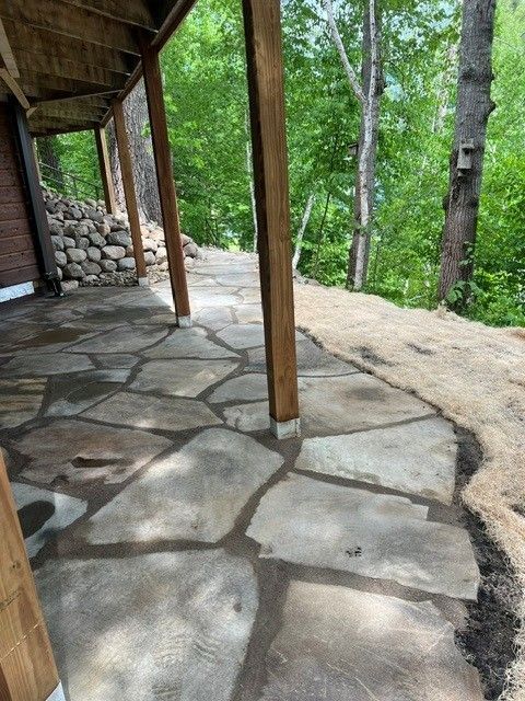 Stone patio under a wooden porch beside a wooded hillside with trees and a stone wall