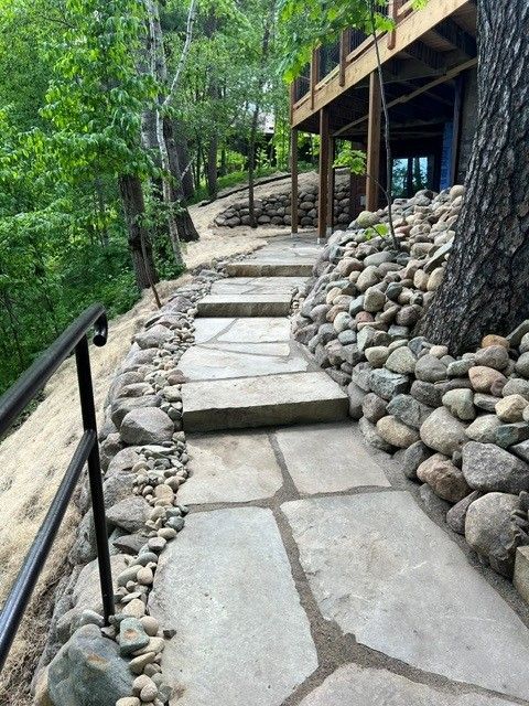 Stone pathway beside a wooded hillside cabin, with railings and stacked rocks along the edge