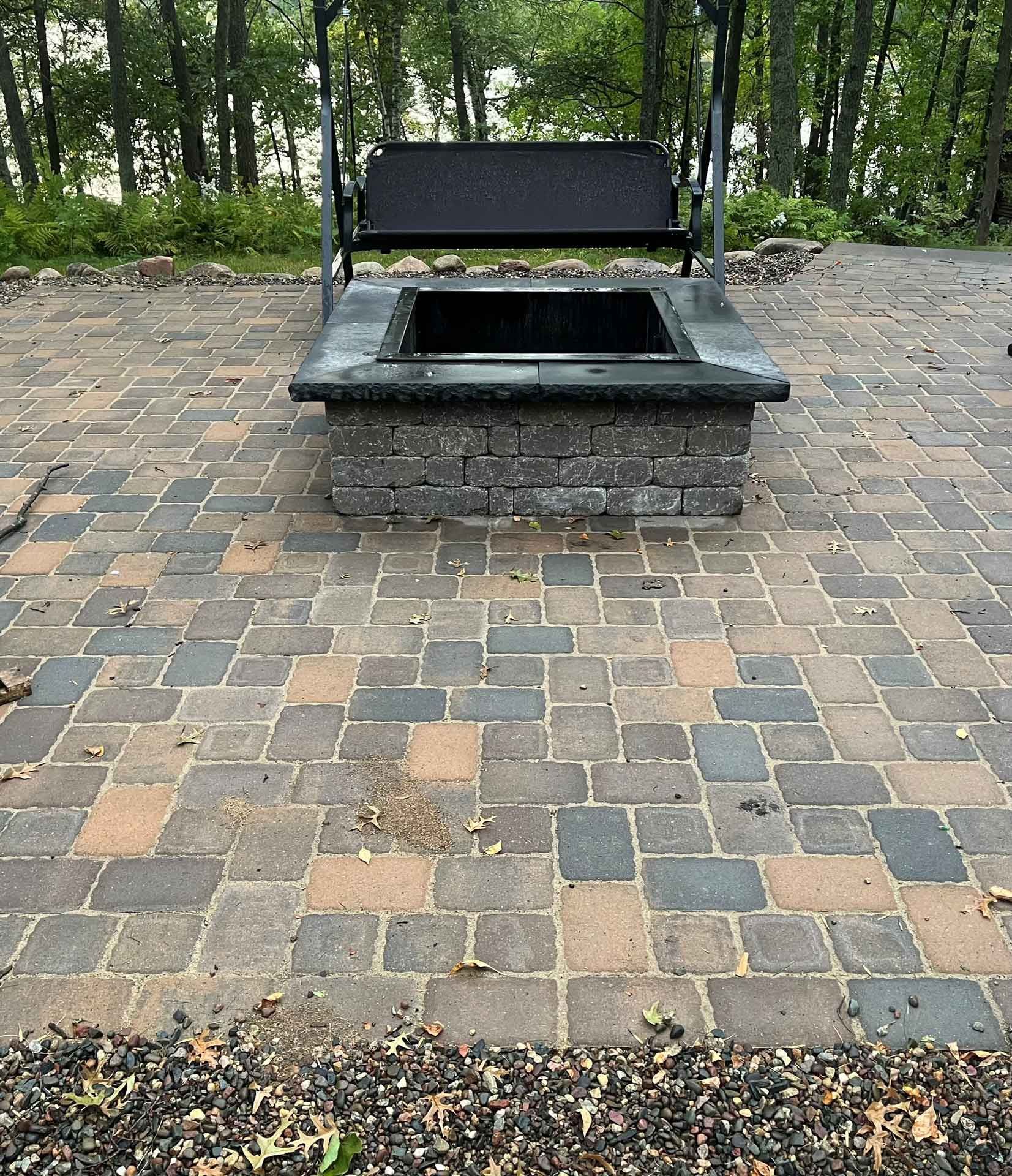 Outdoor brick patio with a black metal bench beside a square stone fire pit in a wooded area