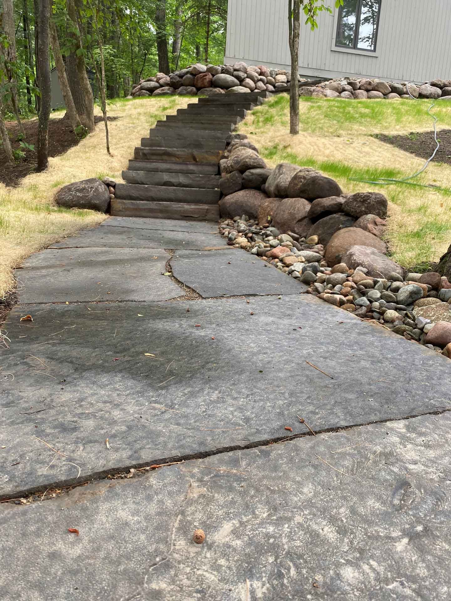 Stone walkway with uneven steps and rocks beside a house in a wooded yard