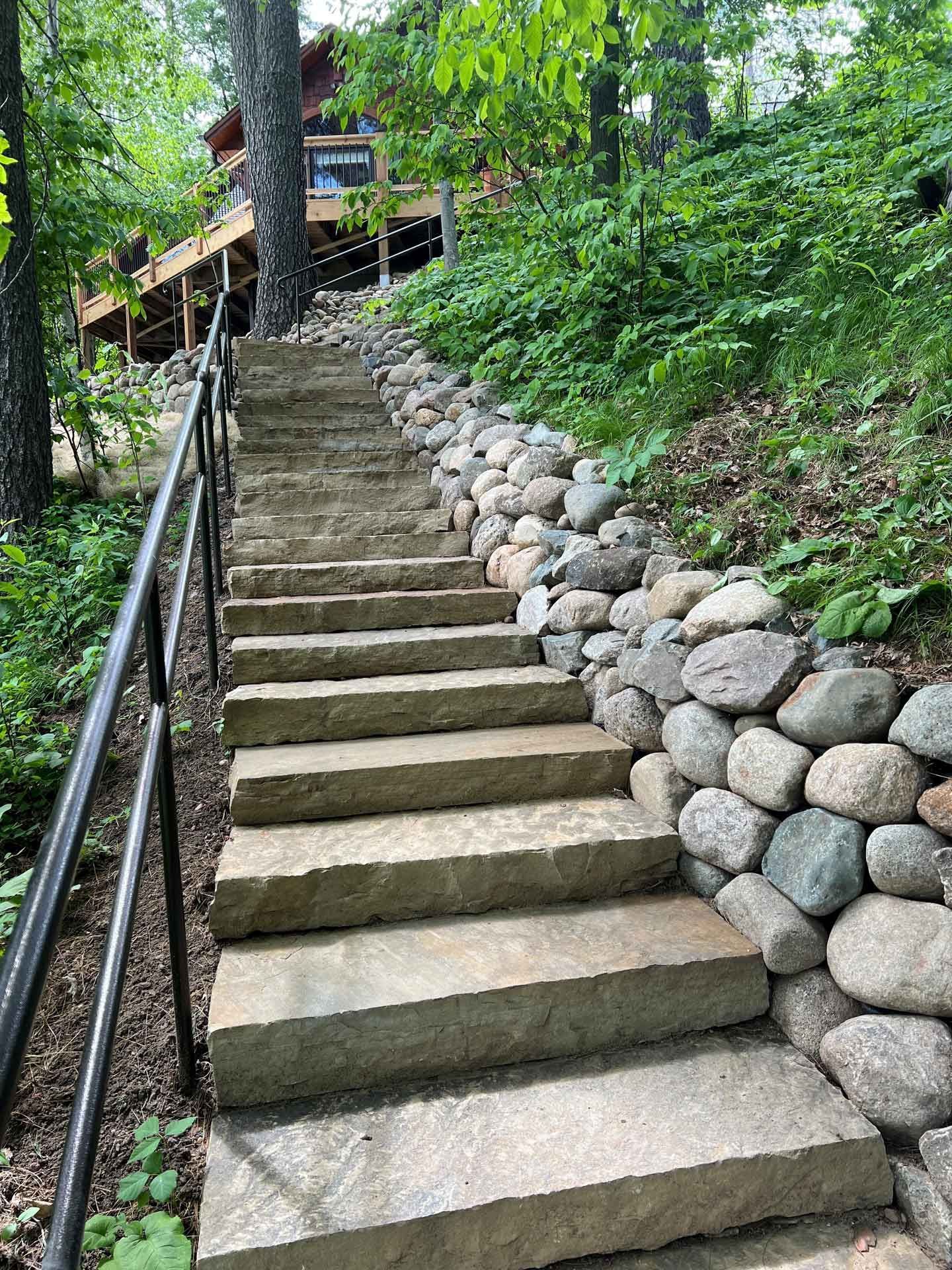 Steep stone staircase beside a wooded hillside, with metal railings and a house at the top.