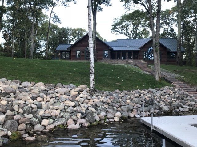 Lakeside log home on a grassy hill, with stone retaining wall and dock. Trees frame the view.