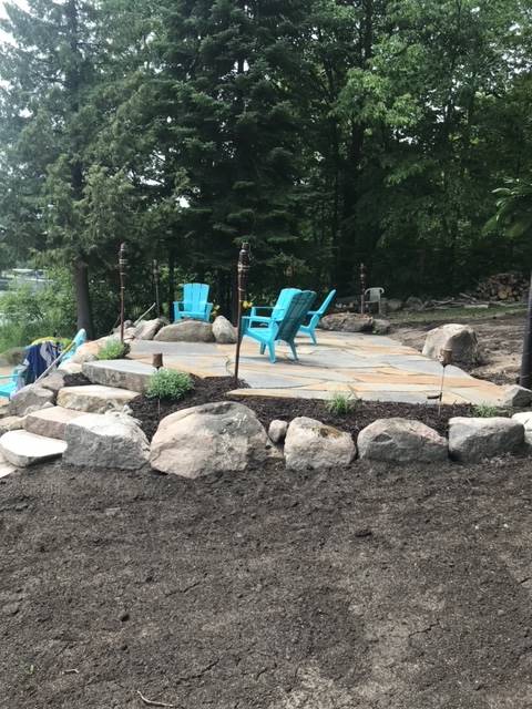 A stone patio with turquoise chairs, surrounded by rocks, dirt, and trees.