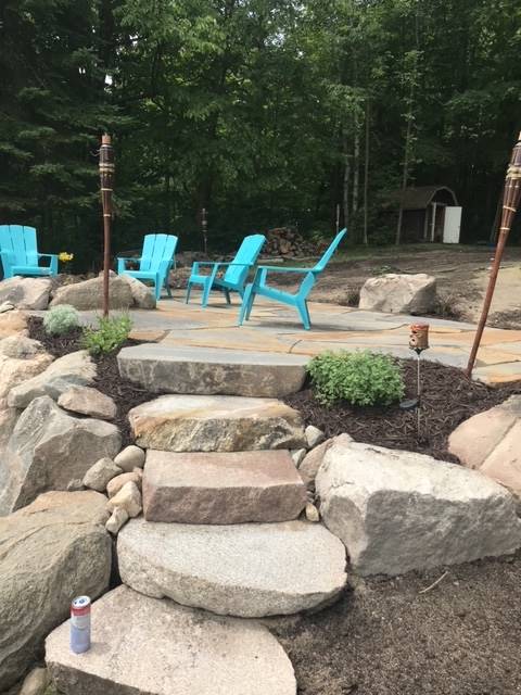 Stone steps lead to a patio with blue chairs, surrounded by rocks and trees.