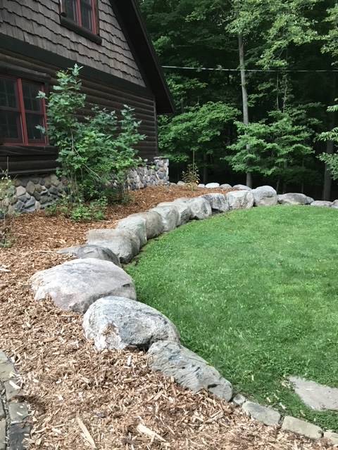 Stone retaining wall separates wood-mulched area from grassy lawn next to a brown-shingled house in a wooded area.
