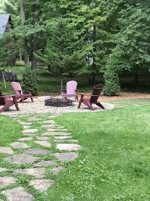 A stone path leads to a fire pit surrounded by Adirondack chairs on a grassy lawn, under trees.